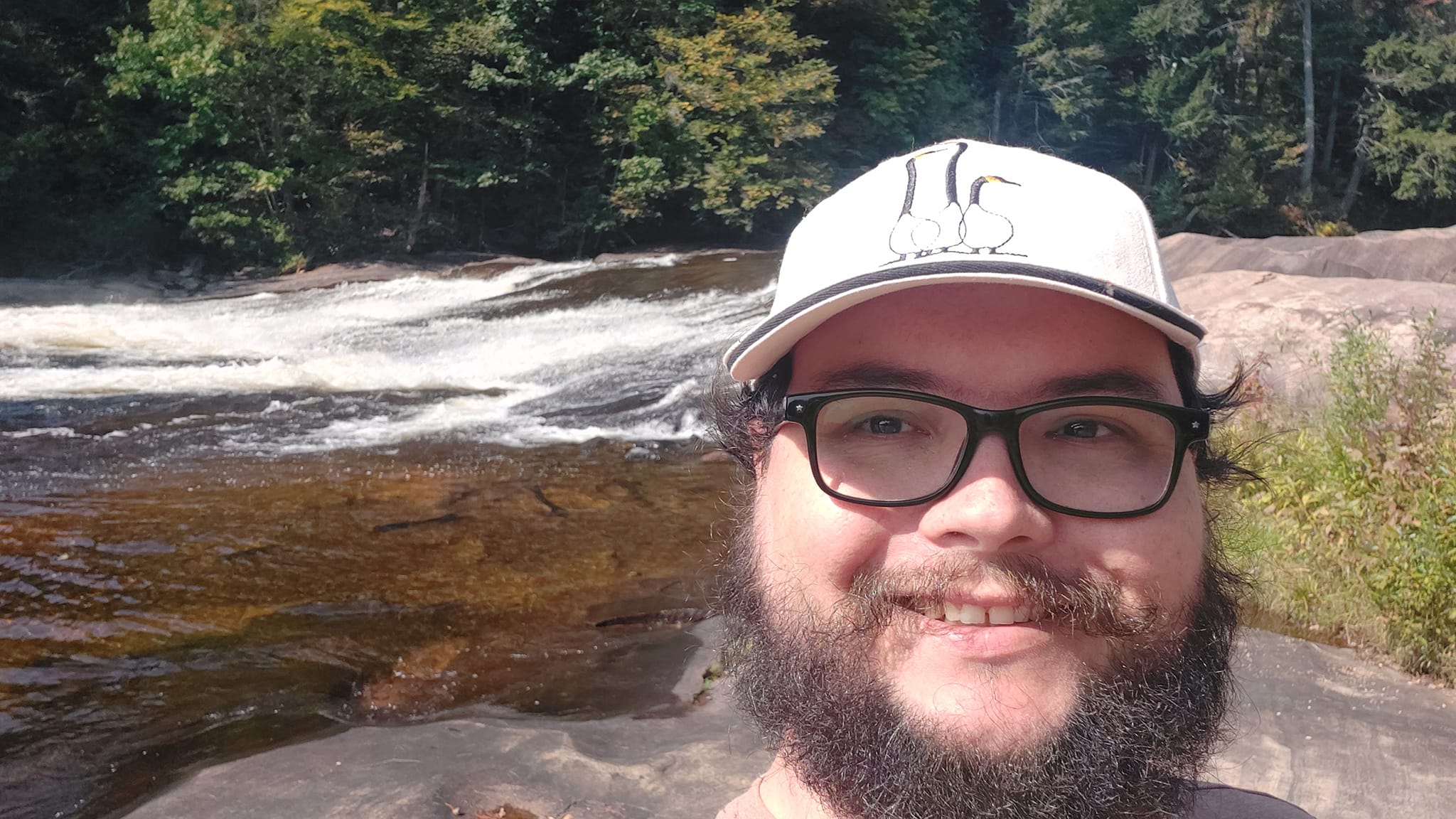 A man with short hair and bushy facial hair stands in front of a low waterfall with no crest, the Canadian shield rocks are identifyable as gneiss from the texture visible in the photo